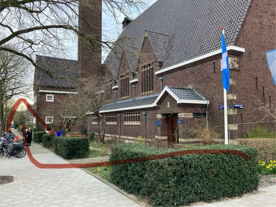 A brown brick church building viewed from the street corner. A red arrow indicates a direction to walk along what is Herman Gorterstraat.