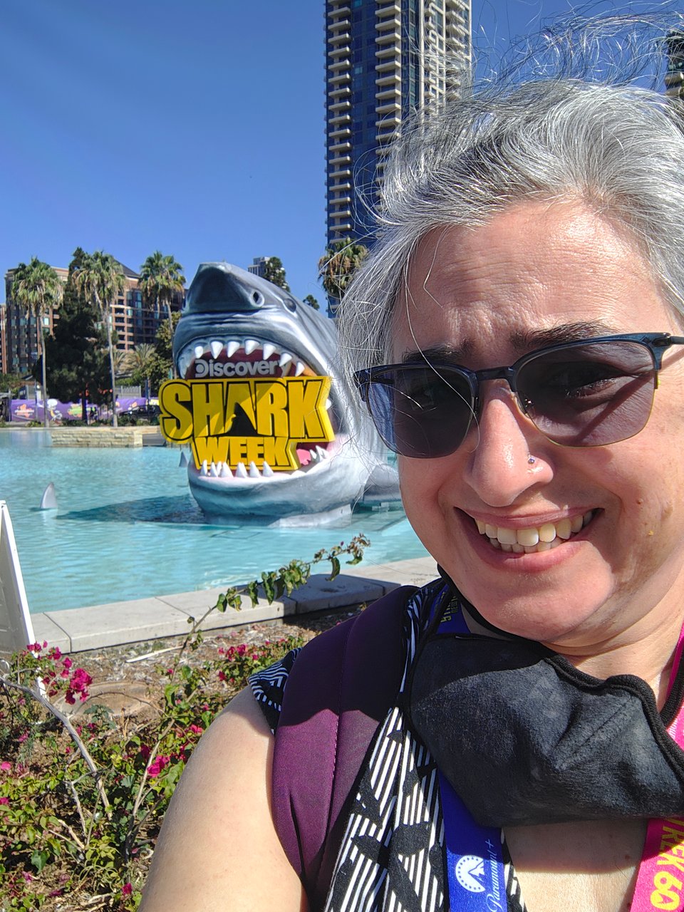 Me, a white woman in a sundress wearing a backpack and ComicCon lanyard smiles with concern outside in a San Diego park, where behind me Discovery has placed a large model of a Great White Shark chomping down on a sign for "Discovery Shark Week" inside a shallow fountain.