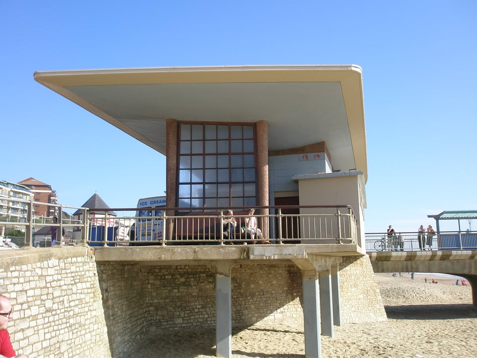 The side elevation of the pier, on a sunny day. The wing roof sits above the shops, which are hidden by a large glass wall with a grid of square panes. Two people are sat on the wooden bench in the shelter.