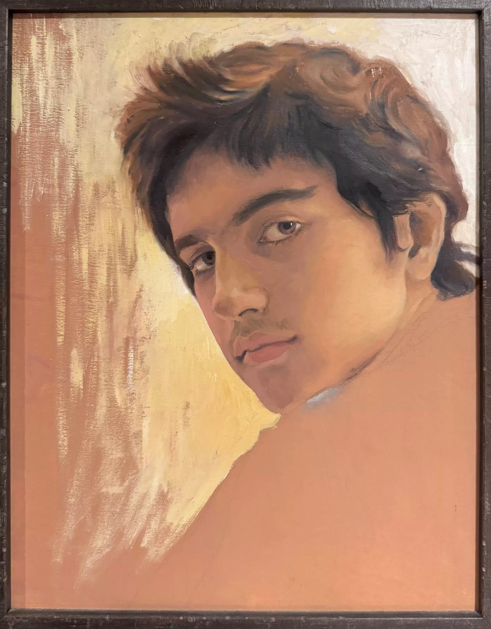 A painting in hues of peach, brown, and buttercup showing Zohran Mamdani at age 14, his face in three-quarter view turning back to regard the viewer over his left shoulder. His hair is slightly unruly, his expression serious, there is a pale yellow glow to his left indicating a light source.