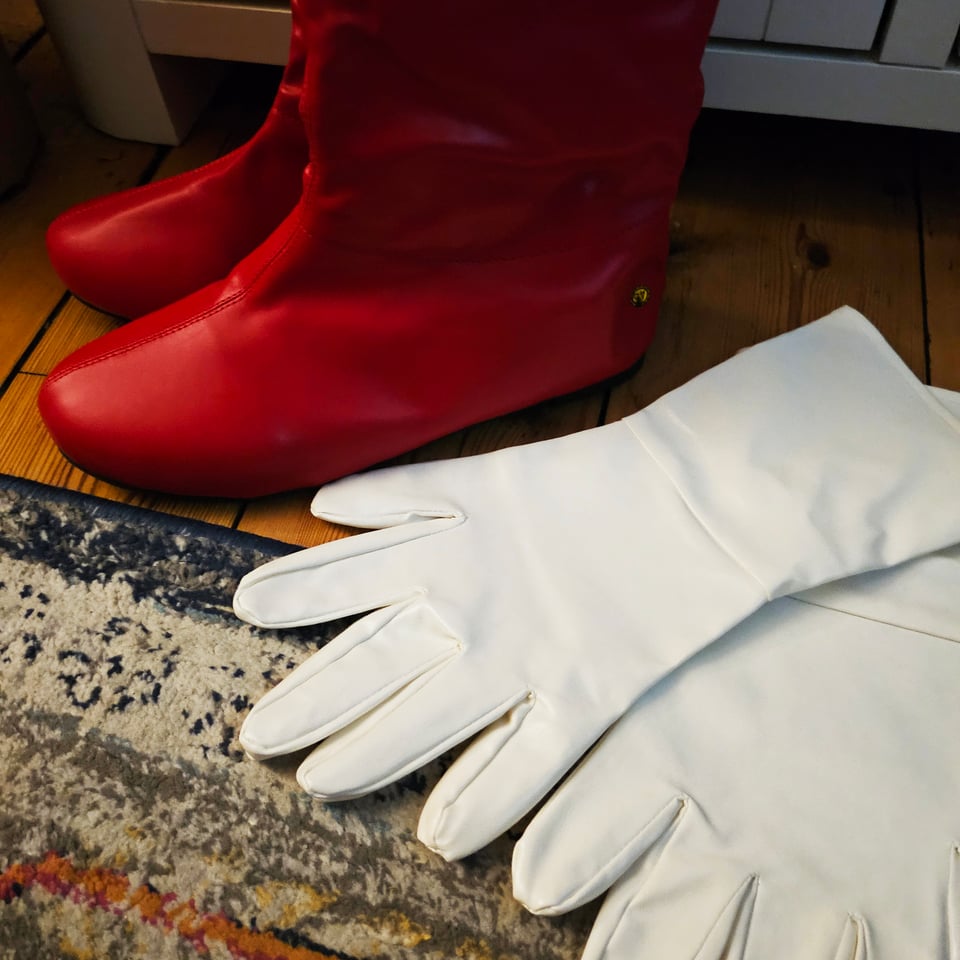 A photograph displaying a pair of red superhero boots and white superhero gloves beside each other on a carpeted floor.