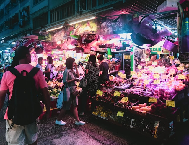 Esta imagem captura a atmosfera vibrante de um mercado de rua noturno em Hong Kong, mergulhado em uma iluminaÃ§Ã£o neon em tons de rosa e roxo. No centro, barracas exibem pilhas organizadas de frutas frescas e vegetais sob toldos coloridos, enquanto pessoas caminham e fazem compras. Em primeiro plano, Ã  esquerda, vemos as costas de um homem usando uma camiseta rosa e uma mochila preta, observando a movimentaÃ§Ã£o. Ao seu lado, uma mulher com um vestido verde e uma bolsa azul caminha em direÃ§Ã£o Ã s mercadorias, enquanto outros clientes interagem com os vendedores ao fundo. A cena Ã© rica em detalhes visuais, desde as etiquetas de preÃ§os amarelas nas frutas atÃ© a arquitetura urbana que se eleva acima do mercado, criando uma sensaÃ§Ã£o de energia e vida cotidiana local.