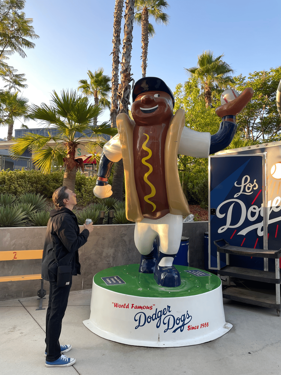 A small human man stares up at a large statue of a hot dog with a face playing baseball