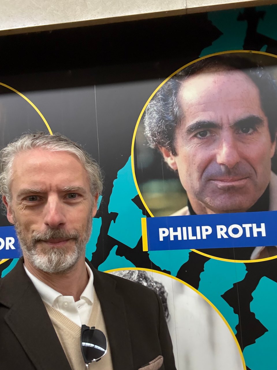 photo of white man in brown cashmere sport coat, light-brown cashmere sweater and white polo shirt, standing in front of a labeled photo of Philip Roth