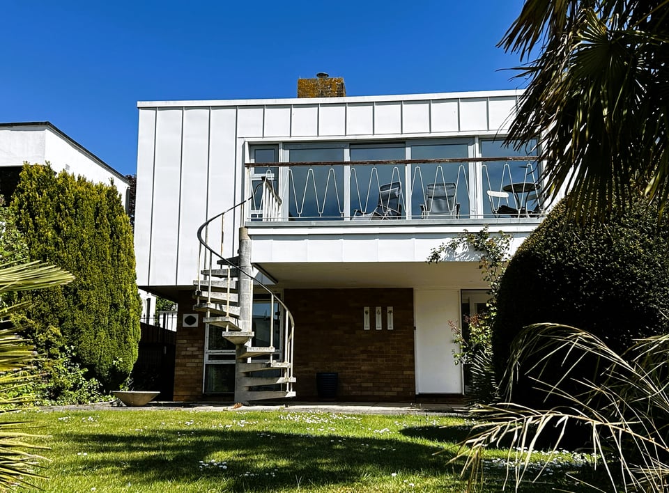 A 1960s upside down house, mostly hidden by mature trees. The upper storey overhangs the ground floor, creating a balcony that can be reached by a concrete spiral staircase. The balcony handrail is wood, and the railings are a 1950s metal wave pattern, painted white. The upper story is now clad in vertical metal panels.