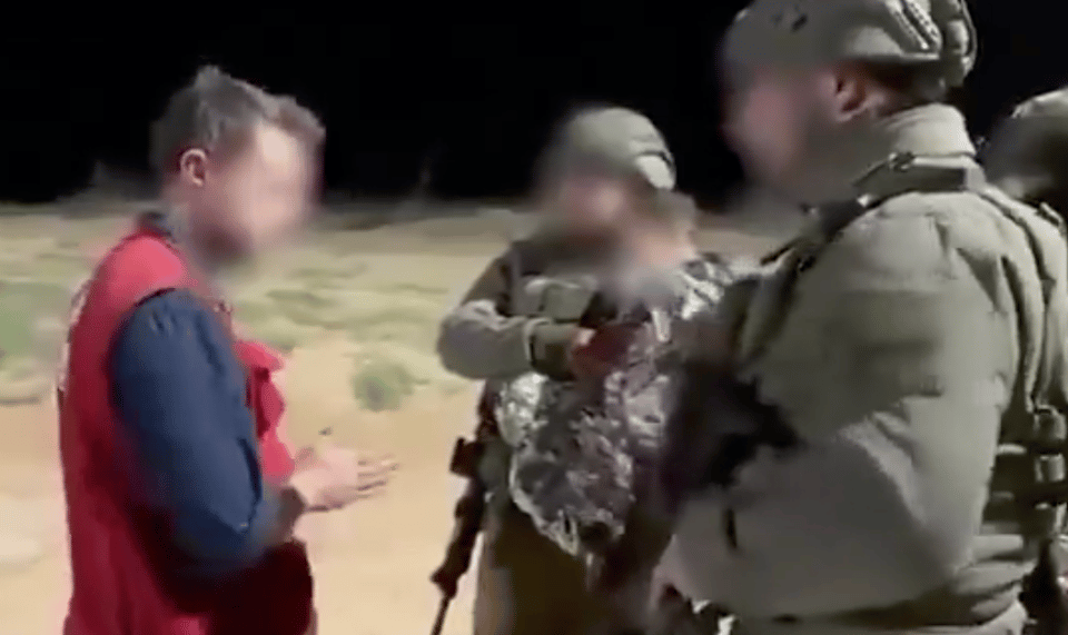 An image of soldiers in helmets carrying guns who are facing a man with a dark shirt and a red vest.  One of the soldiers is carrying what looks like a child in a blanket.  All faces are blurred