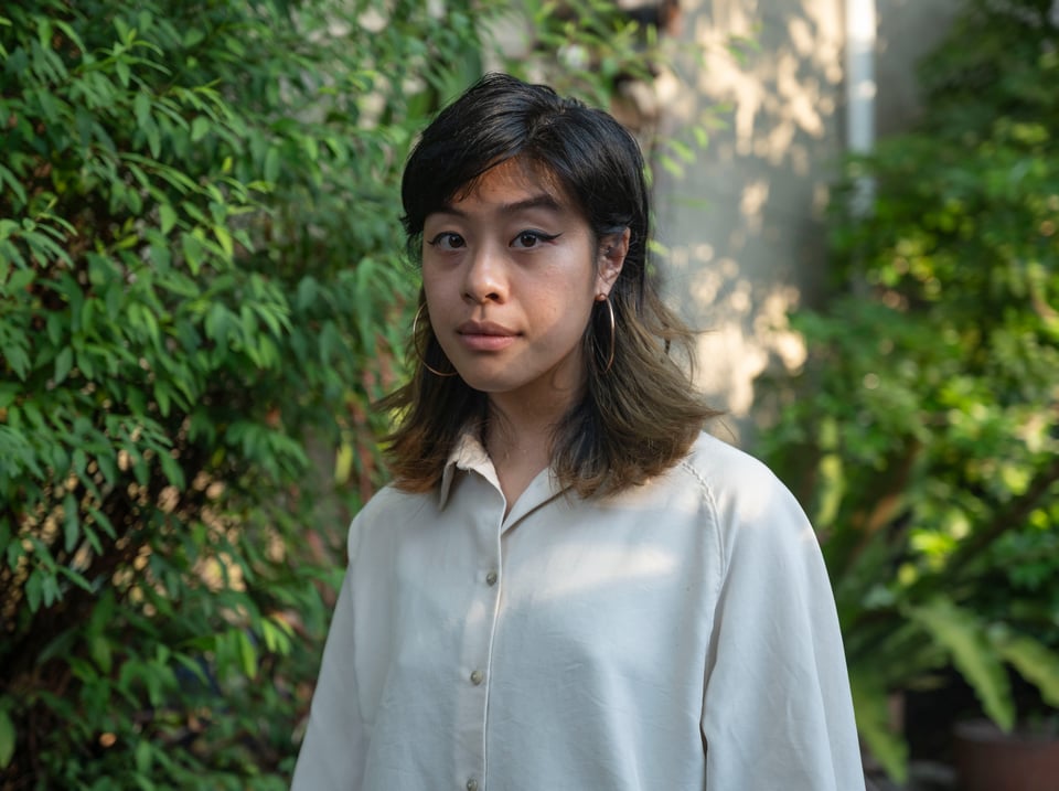 an asian woman in a loose off-white button down shirt looks at the camera, behind her are green plants, light shines on her face