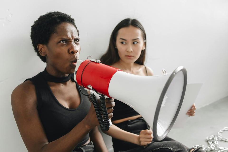A woman with a determined expression speaks into a red and white megaphone, her intensity contrasting with the neutral white wall behind her. Next to her, another woman with a calm but curious expression looks on, holding a piece of paper. Chains are visible on the floor, adding a sense of drama or activism to the scene.