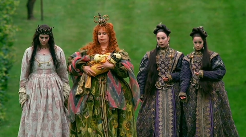four women in heavy gowns, three of them dark haired, one of them red haired, walk along brilliant green grass. The red haired woman is carrying a baby wrapped in yellow cloth.