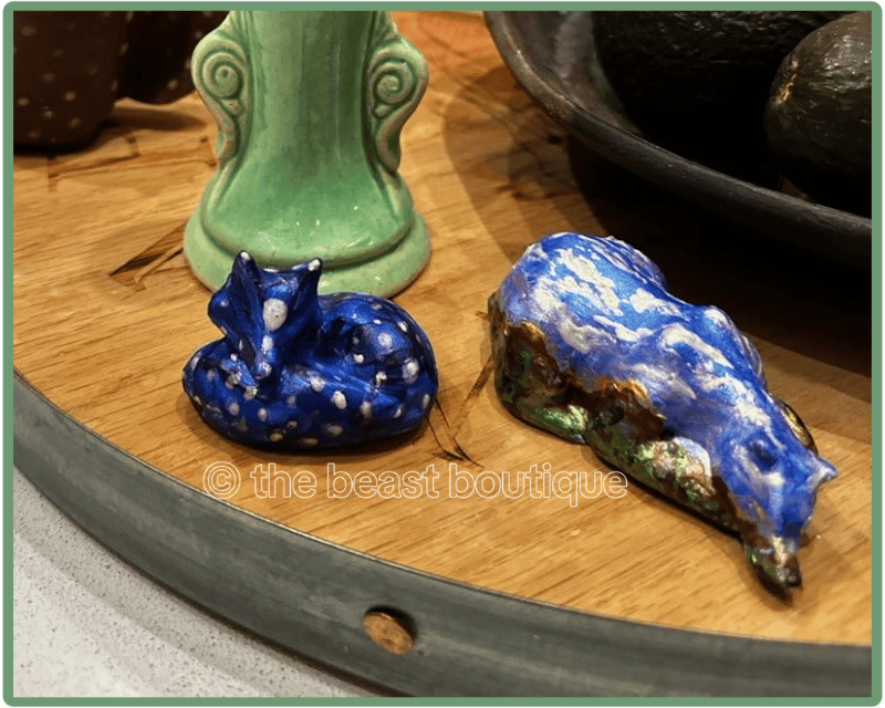 A photo of two figurines, a sleeping fox and sleeping bear, resting on a wooden table. The fox is painted dark blue with silver polka dots on it. The bear is painted in metallic colors like a mountainous desert landscape with a brilliant blue sky.

the shop name, the beast boutique, watermarks the image.