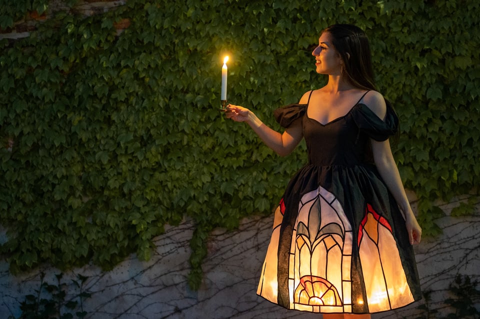 A woman stands in front of a lush wall of ivy at dusk, holding a lit candle in her outstretched hand. She wears a dramatic black dress with puffed sleeves and a glowing, illuminated skirt. The skirt is designed to resemble stained glass, featuring bold, arched patterns in red, yellow, and pale pink, backlit to create the effect of light shining through a cathedral window. She looks like a character from a Disney storybook