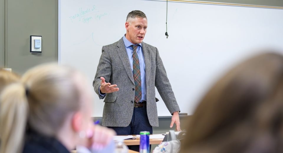 JMC Prof. Tobin Craig wearing a grey blazer and checked tie, teaching in a Case Hall seminar room.