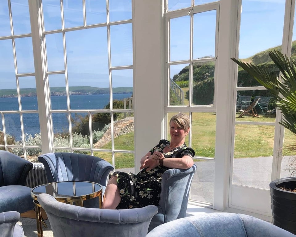 A white woman with short grey hair sitting in a blue velvet bucket chair in a palm court. Behind her is a wall of glass in square panes and the view across to the Devon coast. It is a sunny day, but the woman is checking her watch.