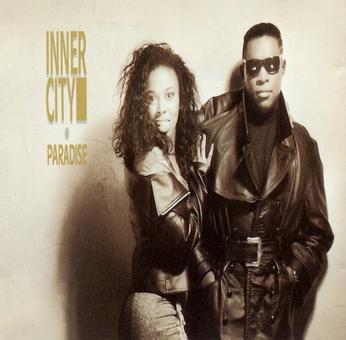 A light-sepia toned photograph of a Black woman and a Black man. They look at the camera. She has long black hair and is turned with her hip towards us and has one arm around his back and the other on his arm. She wears a leather jacket. She has a look of surprise on her face. He has short black hair, sunglasses, and wears a long black leather trenchcoat, with a black shirt underneath. He looks stoic. It is a very MATRIX look, which wouldn't come out for another 10 years.