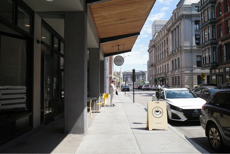 Street-level view of retails storefronts with a wood-paneled overhang and outdoor seating on the sidewalk.