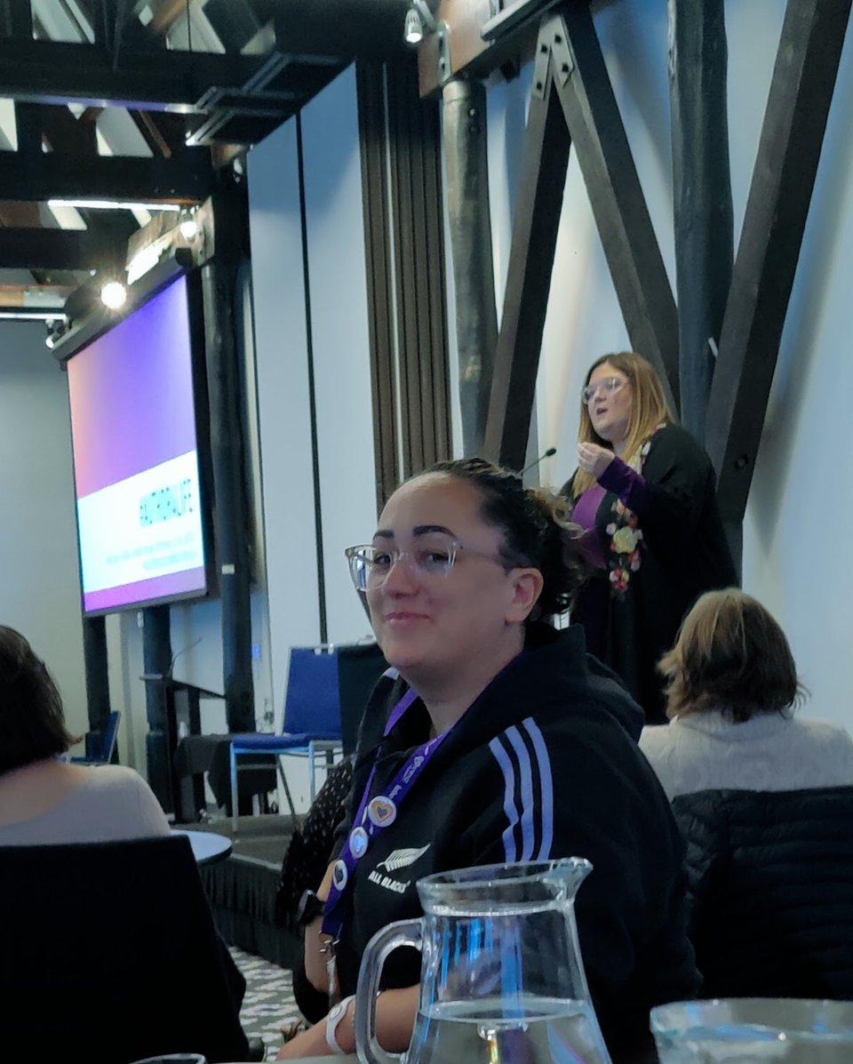 Sacha Black looking cheekily at the camera as Becca Syme speaks from a podium behind her.