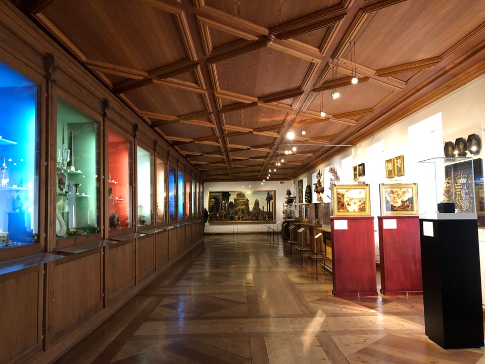 A long, narrow, wooden-floored room with glass cases on either side. On the far wall is a rectangular, painting (landscape orientation). The cases on the left are built into a wooden wall; each is lit in a different colour: blue, red, green. The cases on the right are freestanding; some take the form of stands with small paintings atop.