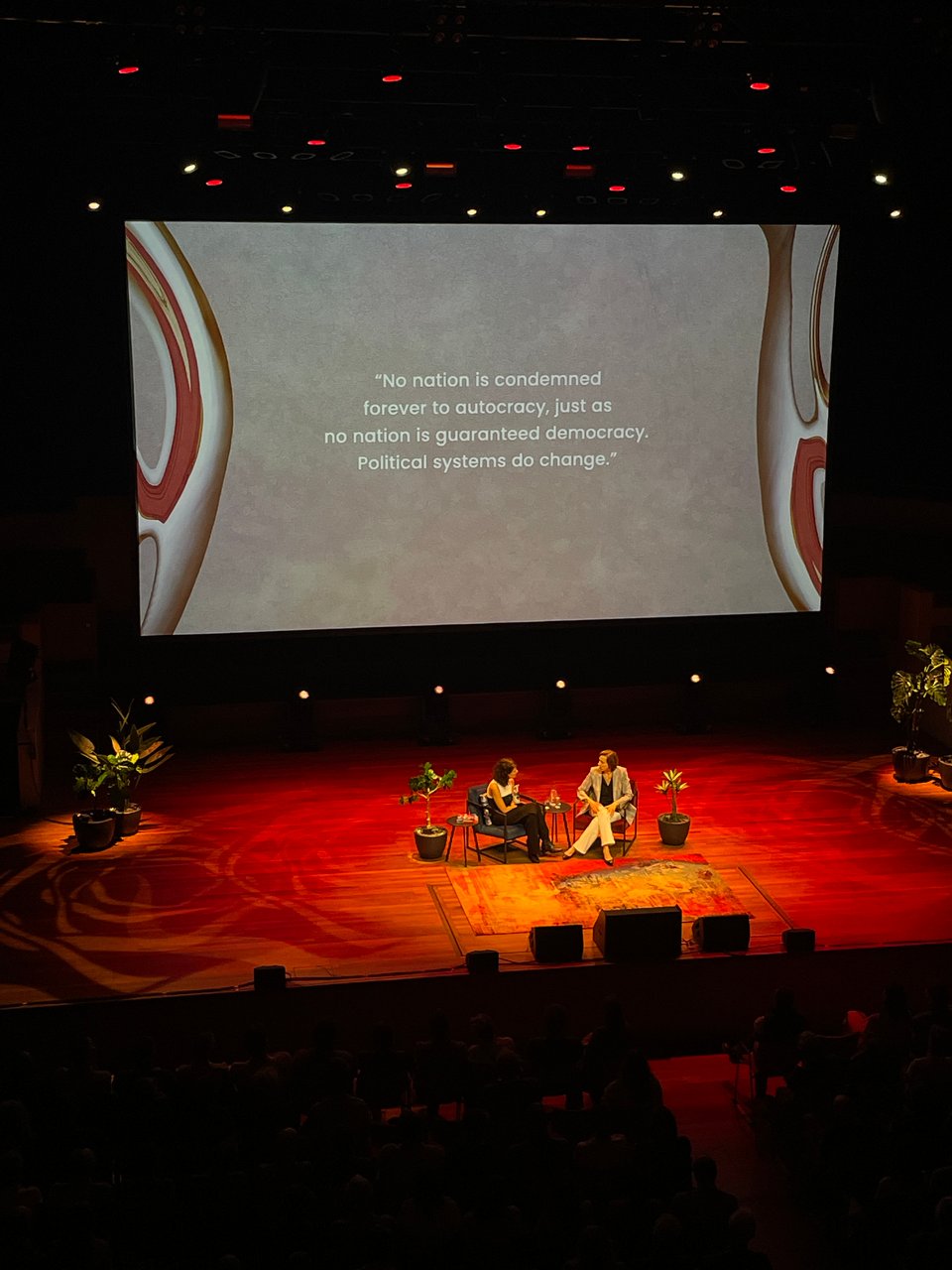 Two women sit in conversation on a stage bathed in red. On the screen behind them it reads: “No nation is condemned forever to autocracy, just as no nation is guaranteed democracy. Political systems do change." by Anne Applebaum