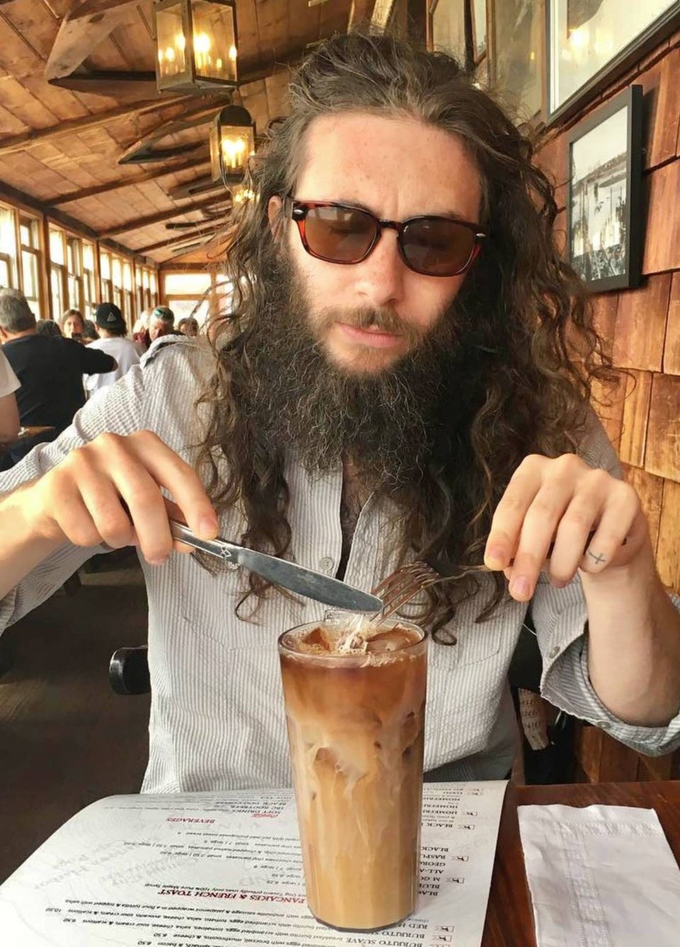 A man with long hair and a bushy beard wearing sunglasses holds a knife and fork poised over tall glass of iced coffee.