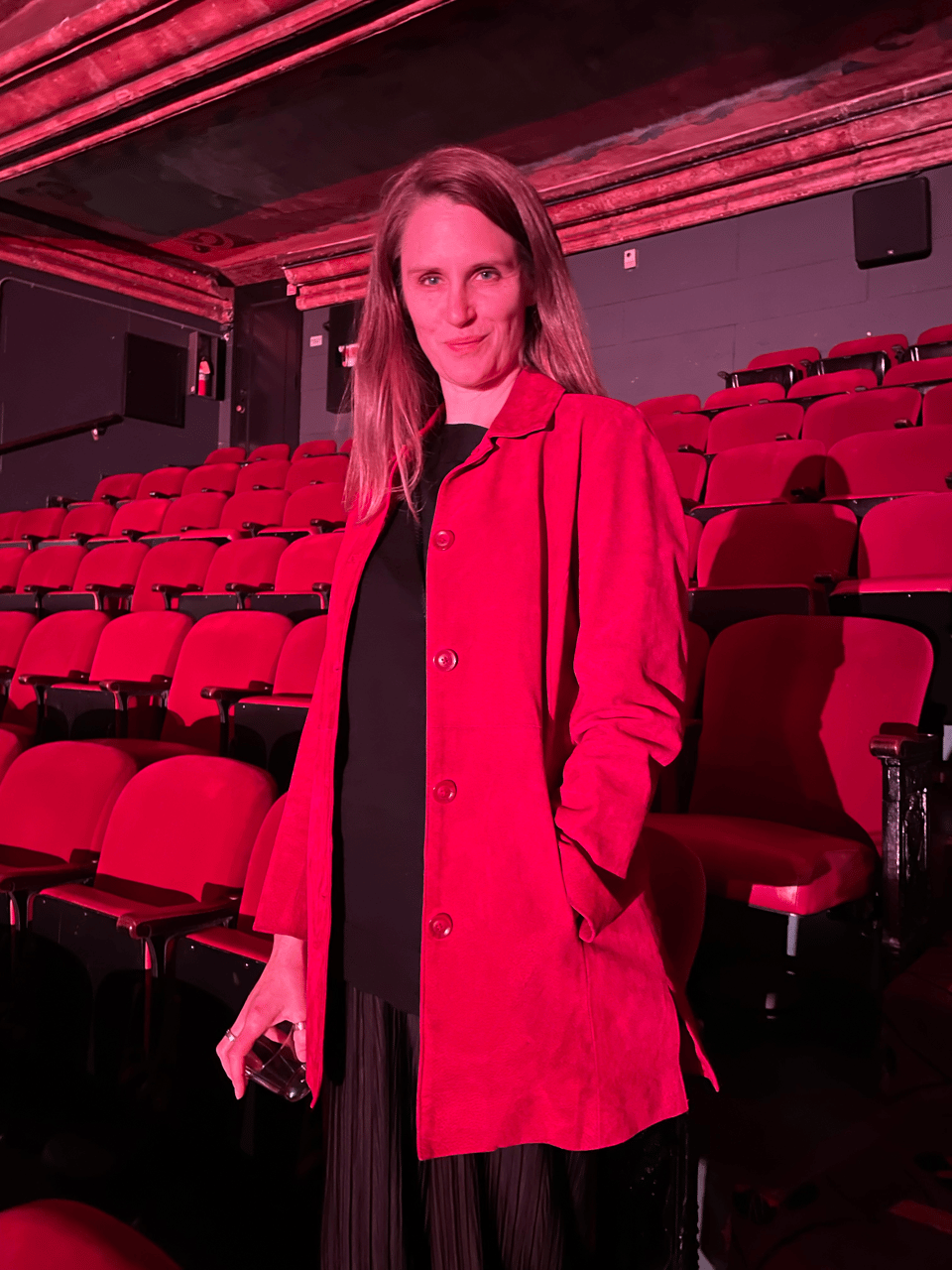 A beautiful woman in a blazing red jacket poses in front of a sea of blazing red theater seats