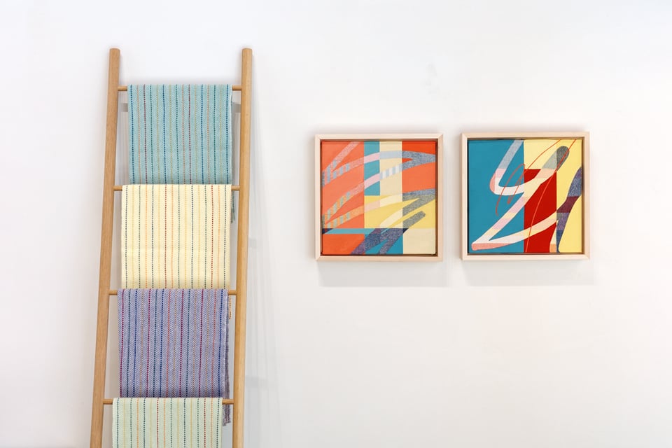 A wooden ladder-style display rack holds four handwoven tea towels in different colors - turquoise at top, cream with thin stripes second, purple/lavender third, and pale green at bottom. To the right, two framed abstract paintings hang on a white wall. The left painting features coral, yellow, and blue geometric shapes, while the right painting displays bold angular forms in teal, red, yellow, and cream tones.