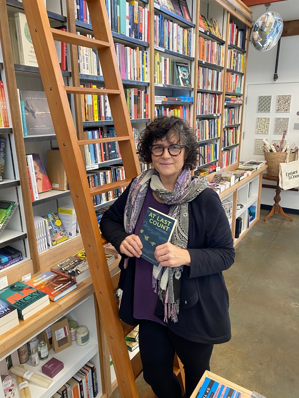 A woman standing in front of a ladder in a bookstore. The woman is author Claire Ross Dunn. She is holding a copy of her novel AT LAST COUNT.