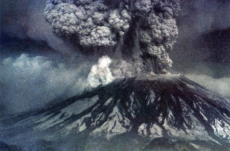 Grainy photograph of Mt St Helens erupting. A plume of ash rises from the crater at the top of the mountain. The image looks like it was taken with color film, but it mostly shades of grey.