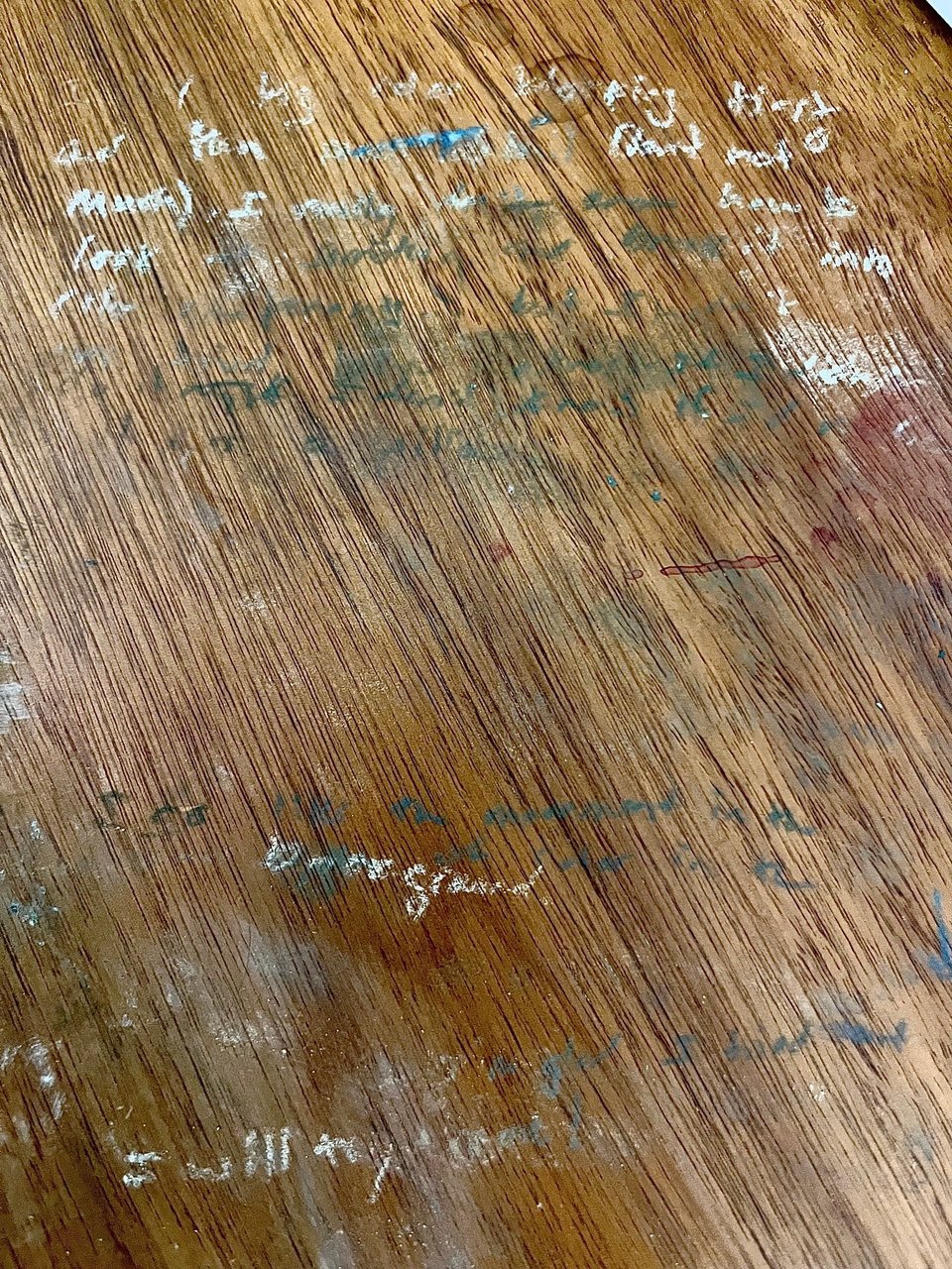 A close up of a dirty wooden desk. Barely legible words in something like chalk are pressed into it in different colors, mostly white. A few phrases are readable: "first try", "background", "I will try..." ("again" is smudged out after that one).