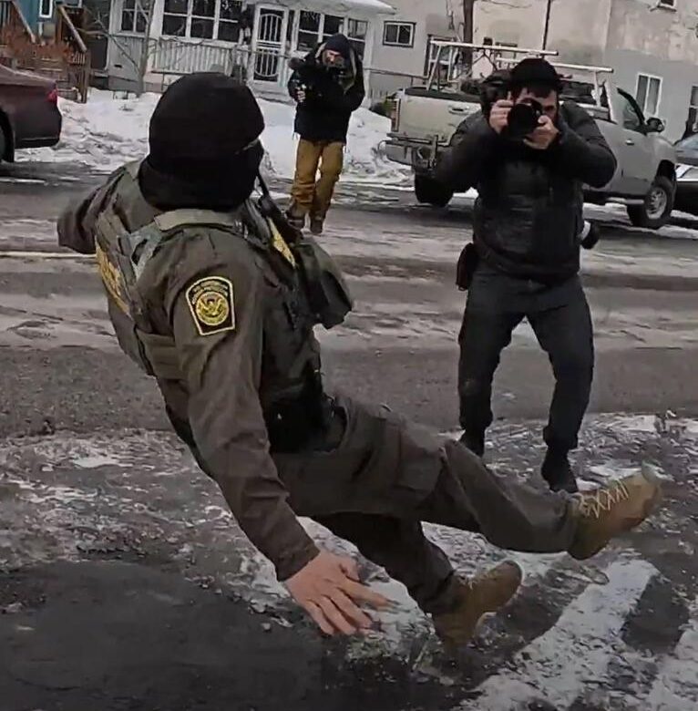 A person snaps a photo as a masked ICE agent slips and pratfalls backwards on ice