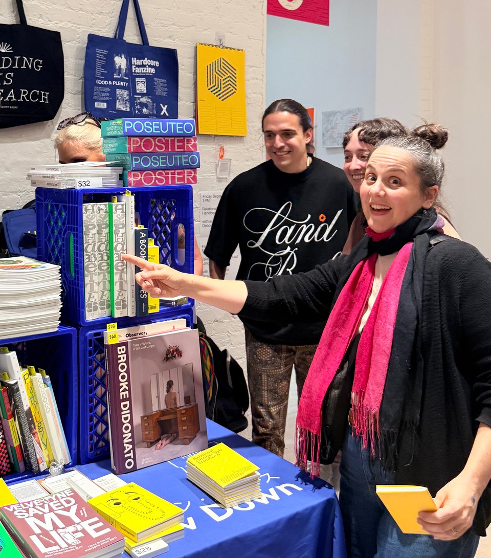 A woman points to book spines at table displaying books. two people stand behind her smiling, there are tote bags and posters displayed on the wall behind the table