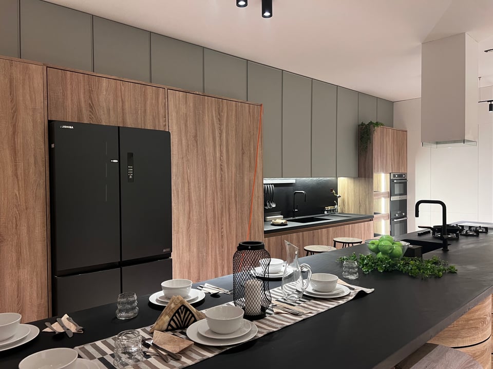 Kitchen cabinets made of light wood, large black refrigerator doors, and an kitchen island with place settings
