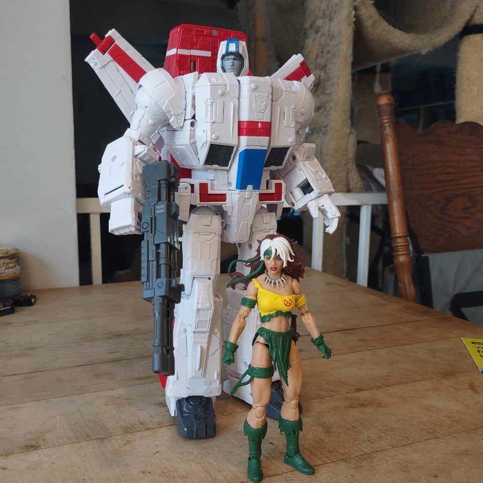 A tall, white and red toy robot (Jetfire of the Transformers) stands on a wooden table behind a (much shorter) action figure of a woman with brown and white hair wearing tattered yellow and green clothes (Rogue of the X-Men, as she appeared in the Savage Land)