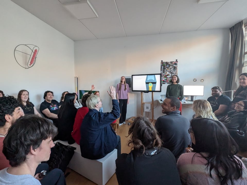A crowded room laughs and reacts to two speakers standing next to a TV that shows a videogame. A man in the center is raising his hand.