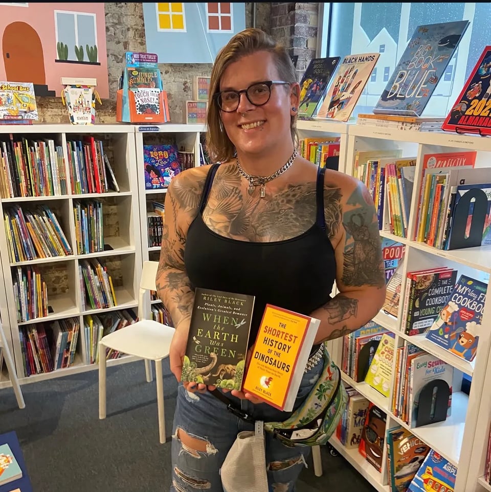Photo of Riley holding a copy of When the Earth Was Green and The Shortest History of the Dinosaurs in Snug Books, Baltimore. They are wearing a black cami and distressed jeans, with a chain-style collar and dark blue glasses. They have many tattoos across their arms and chest, mostly fossils and snarling animals.