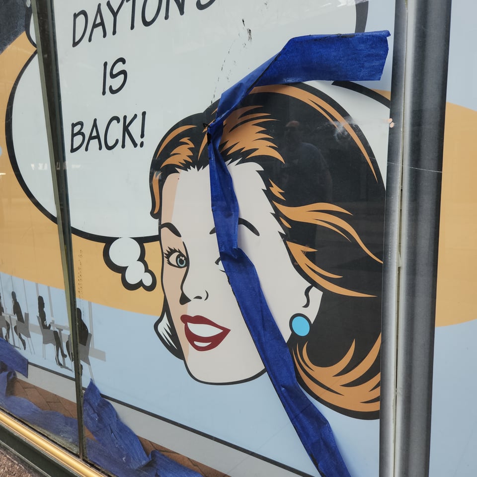 A street-level department store window in downtown Minneapolis. There is a cartoon image of a woman's head with a speech bubble and the text "Dayton's is back!". Strips of blue painter's tape cover part of the woman's face.