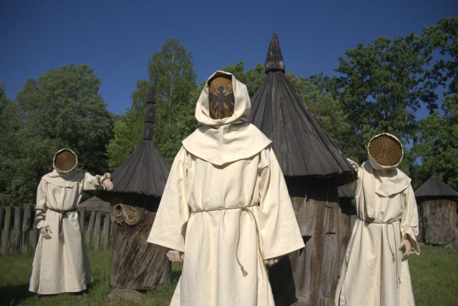 Three men dressed in white cloaks and ropes around their waist. They wear the top of wicker baskets as masks, emulating the outfit of medieval beekeepers. The man in the center has a bee drawn in the mask. They stand in front of straw tents in a green forest.