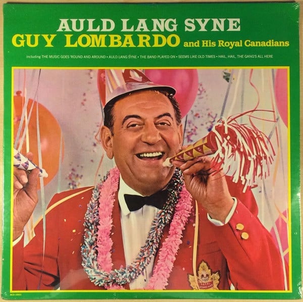 Colour photo of a vinyl record cover. A man wearing a party hat and holding noise makers in his hand with balloons in the background. The man is wearing a red blazer with a maple leaf on the breast, a white collared shirt and a bow tie