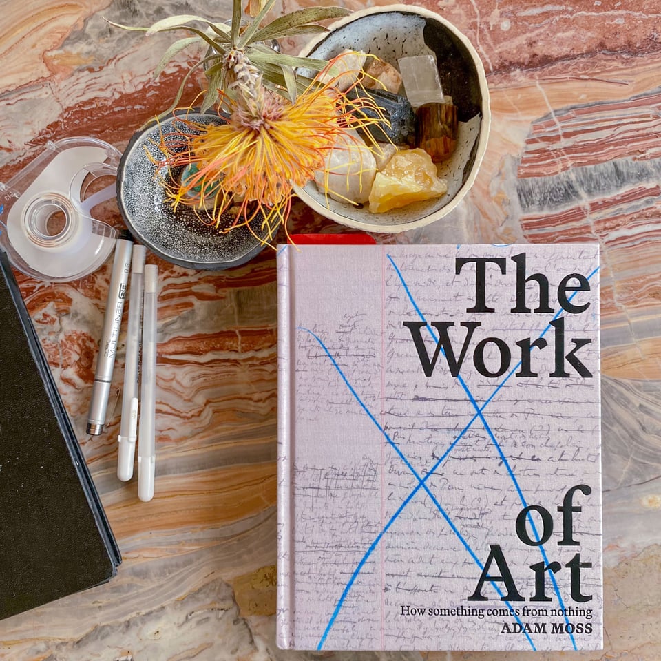 A copy of the book "The Work of Art" on a marble table with some pens, a notebook, a flower and a bowl of rocks.