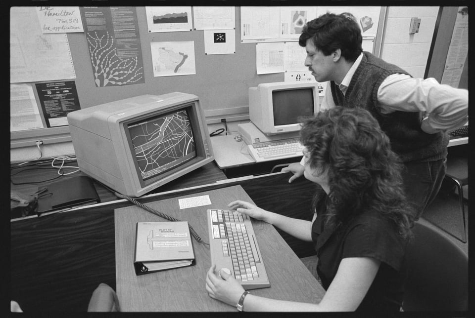 A black and white photograph of a woman sitting at a computer keyboard, looking at a mapping application on a CRT monitor, while a man stands next to her and looks on. There is other errata scattered around them lie a binder, another computer, and a bulletin board with notices pinned to it.
