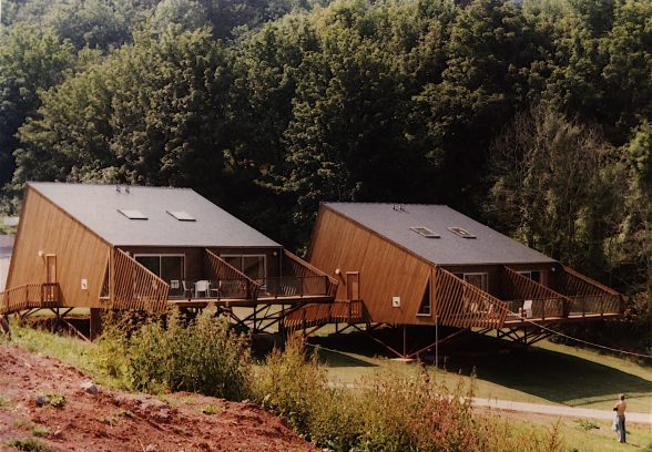 Two pristine pairs of lodges, photographed from the building site of the next ones. Each lodge is a clear wedge shape, with cantilevered balconies. The balconies' glass safety rail is still clear, and almost invisible. They are resting on a grass lawn, with trees behind.
