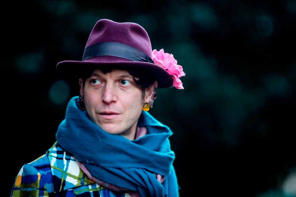 Photo of Mattilda Bernstein Sycamore, a trans femme who wears a colorful jacket, deep blue scarf, and purple hat with a pink fabric flower attached to the right side of the band. She is looking to the left at something out of frame.