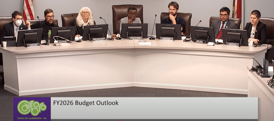 Members of the Carrboro Town Council sit at the dais in the council chambers at Carrboro Town Hall. A chyron at the bottom has the Carrboro logo and the text "FY2026 Budget Outlook"