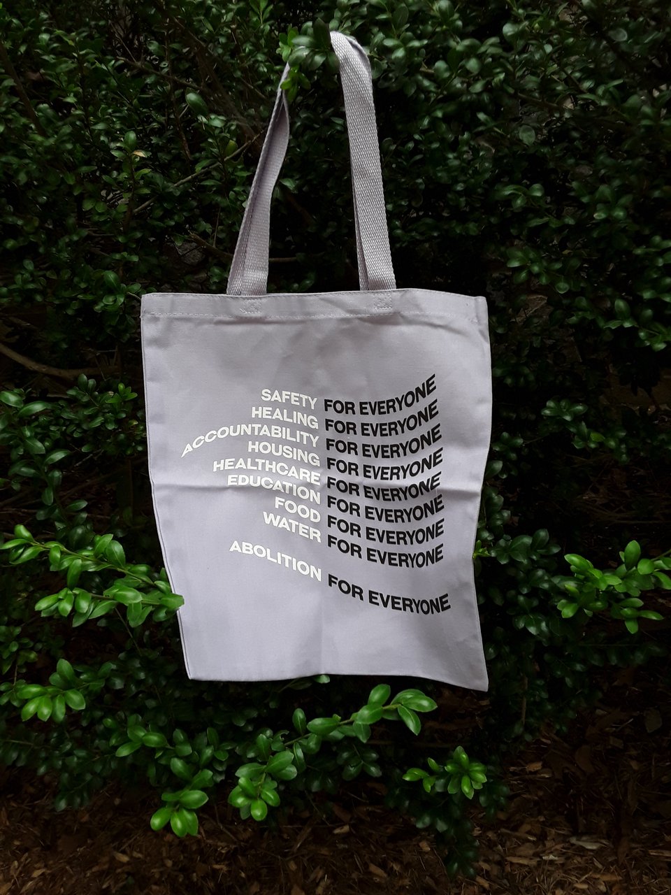 a lavender tote bag in a green shrub in white curved text a series of demands (safety, healing, accountability, housing, healthcare, education, food, water) and then in black after every demand for everyone, on the last line abolition for everyone