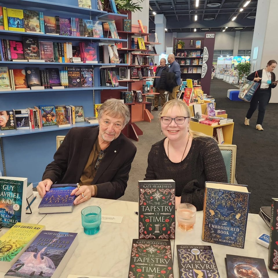 Guy Gavriel Kay and Kate Heartfield sit beside each other at a signing table at the Salon du livre with copies of their books on display in front of them. Kay is leaning closer to Kate for the shot.