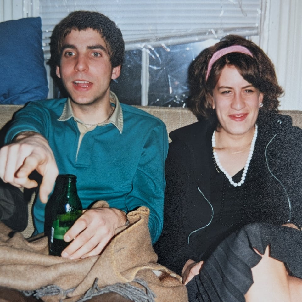 Two early 20 somethings sit on a used couch. The boy is holding a green beer bottle and is pointing past the camera. The girl is wearing a pink headband, black zipper hoodie and a grey pleated skirt. Both are smiling.