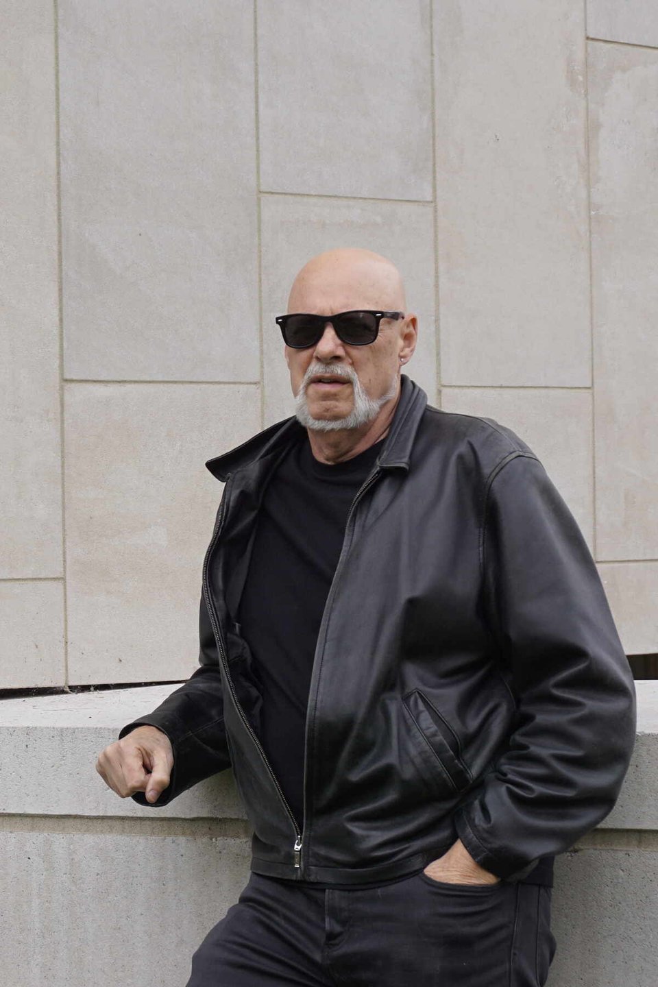 A bald man with a stylized white goatee and wearing dark sunglasses, black jeans a black t-shirt and a black leather jacket stands in front of a concerte wall.