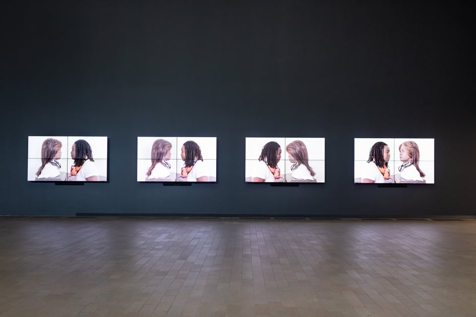 A black gallery wall with four large monitors. Each monitor shows Stephanie and Bina48, face to face, as if in close conversation.