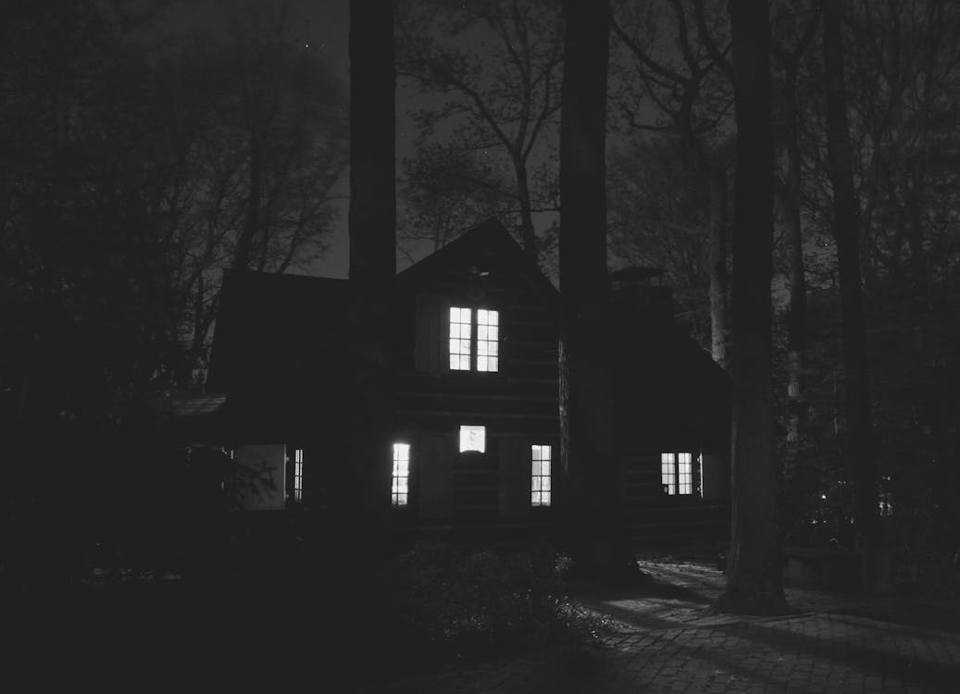 A dark, moody photograph of a house at night shows a cabin-like structure surrounded by tall trees. The building’s windows glow from within. It's pretty creepy