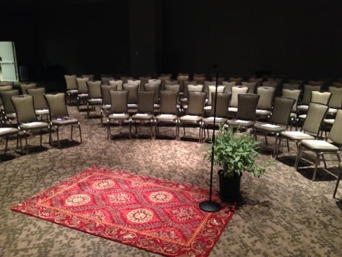 Photograph of a large conference room. In the center is an Oriental rub with a single microphone and a potted plant. Rows of chairs surround it in an oval shape.