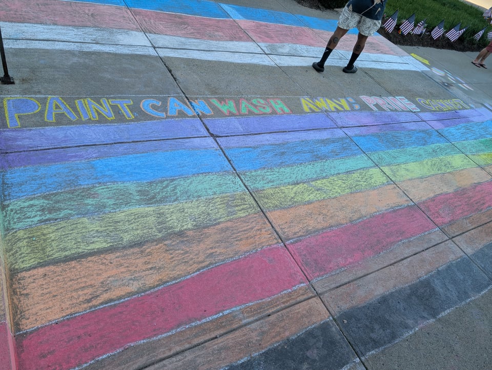Chalked flags on the sidewalk. Pride flag on the bottom of the image, Trans flag in the top of the image. In between is written "Paint can wash away; Pride cannot"